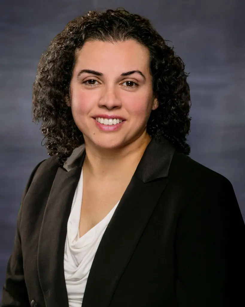 A woman with curly hair wearing a black suit and white top, smiling at the camera.