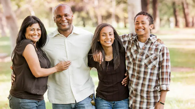 A family of four posing together outdoors in a park setting, smiling at the camera.