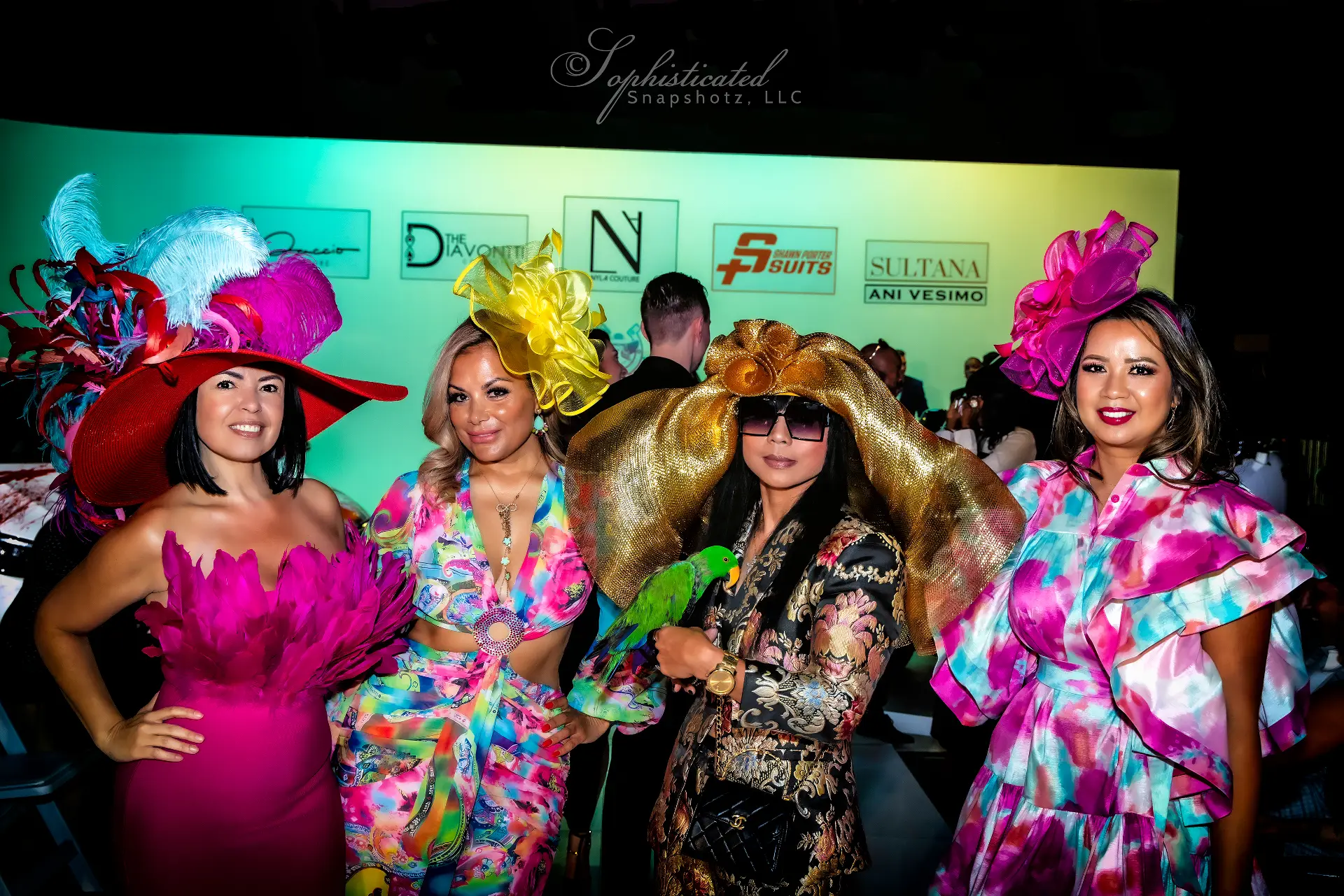 Four women wearing vibrant hats and outfits, posing together at a social event with a colorful backdrop.
