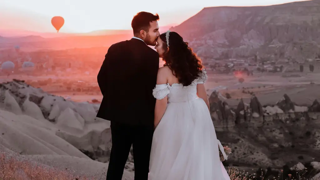 A couple in wedding attire standing together with a sunset backdrop and a hot air balloon in the distance.