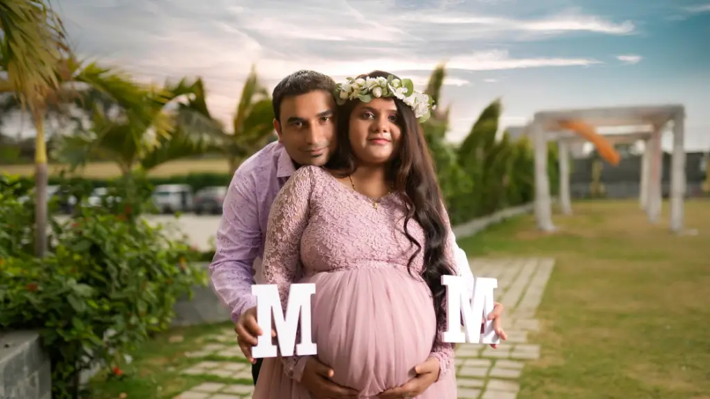 Pregnant woman in a pink dress holding letter blocks with her partner behind her, both smiling.