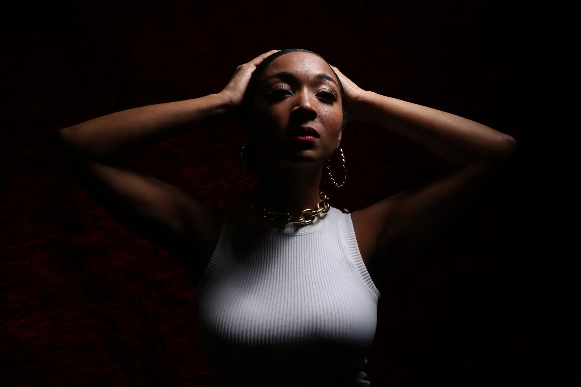 A woman with her hands on her head, wearing a white top and large hoop earrings, in low light conditions.