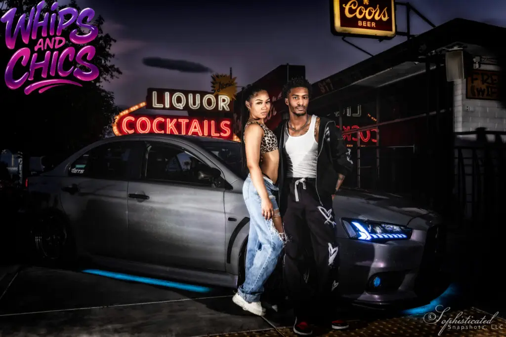 A couple stands next to a silver car in front of a neon-lit liquor store sign at night.